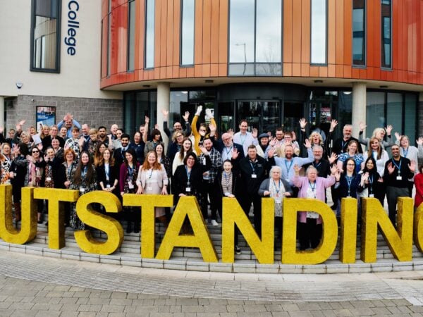 Smiley crowd of people stood with arms in the air in celebration behind large gold sparkly letters that read 'Outstanding'.