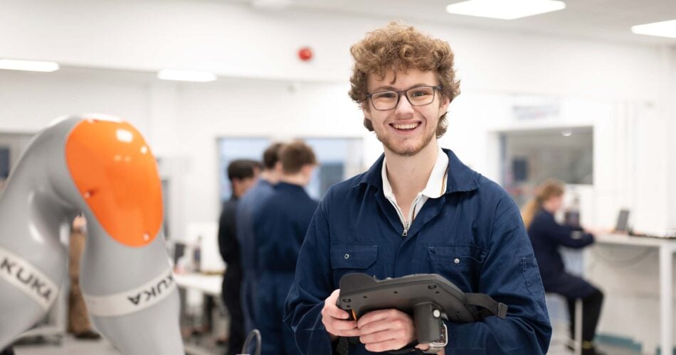 A smiling student with a tablet, next to a robot arm
