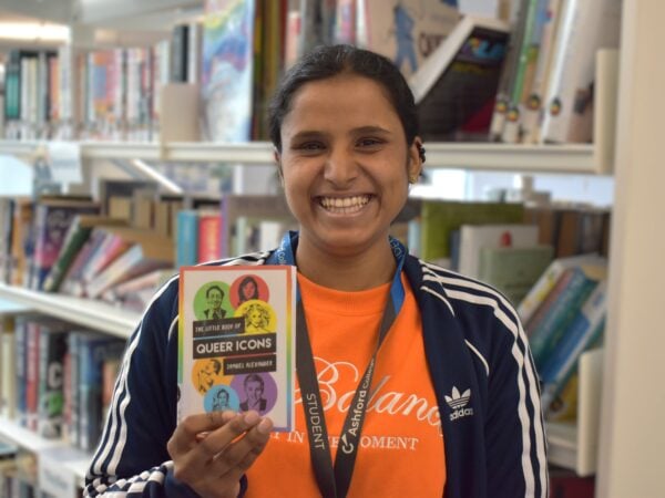 Student stood in a library holding a book titled 'The Little Book of Queer Icons'
