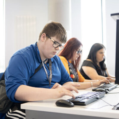 three students sitting at desks working on laptops. Student nearest is wearing glasses and a dark blue shirt,