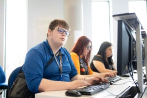 three students sitting together at computers. Closest student is looking at the screen, they are wearing glasses and a dark blue shirt