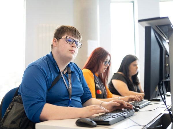 three students sitting together at computers. Closest student is looking at the screen, they are wearing glasses and a dark blue shirt