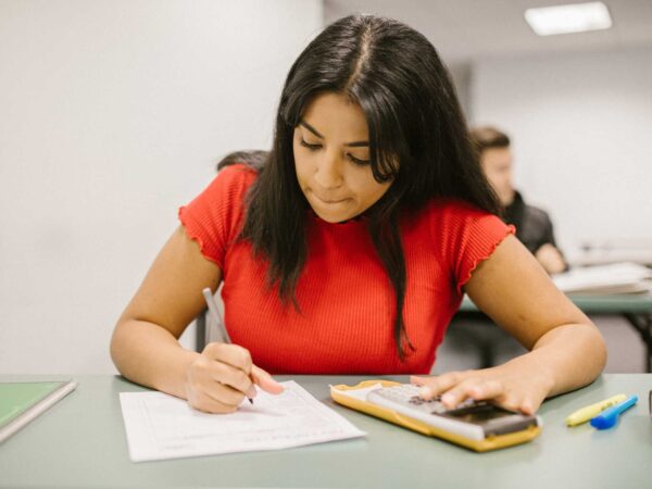 Student in a red top with long black hair sitting at a desk writing with a pen onto white paper and has a calculator.