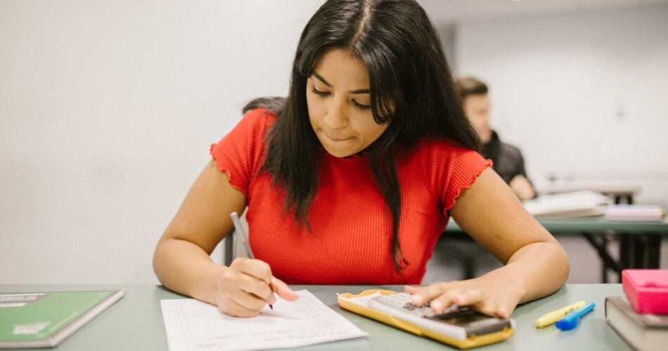 Student in a red top with long black hair sitting at a desk writing with a pen onto white paper and has a calculator.