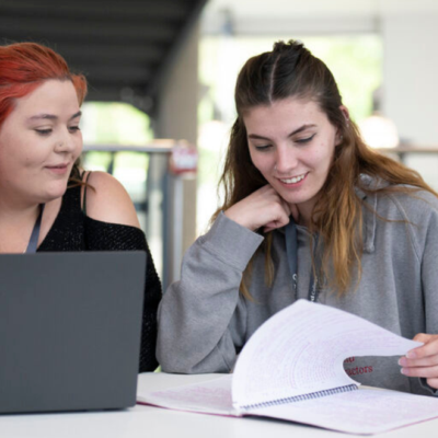 two students sitting at a desk, one student has a laptop and is looking at the other student who is looking through a study book.