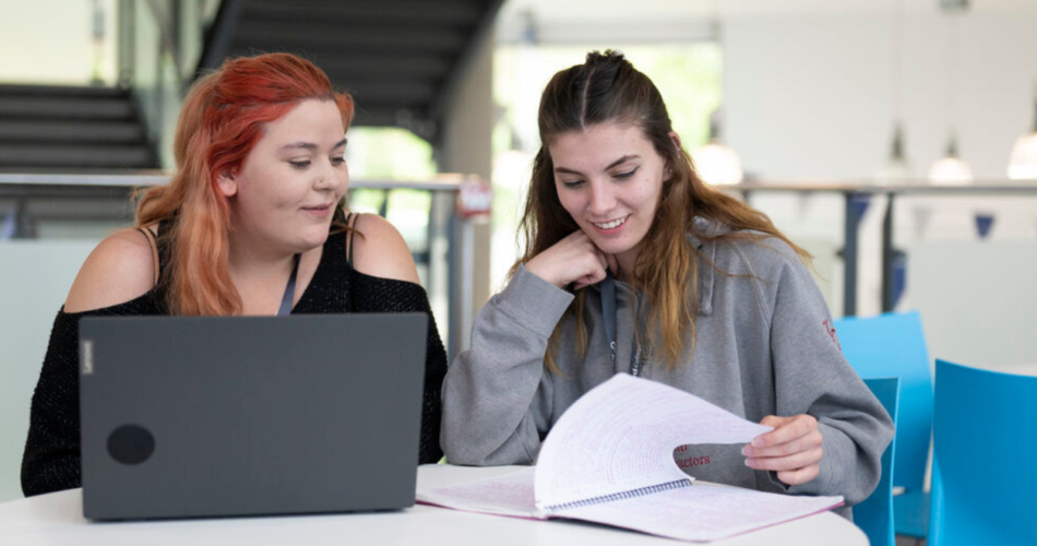two students sitting at a desk, one student has a laptop and is looking at the other student who is looking through a study book.