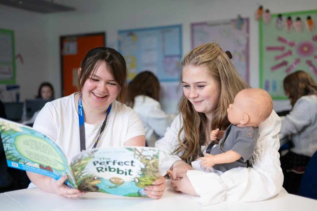 two students reading a book at a table to a doll.