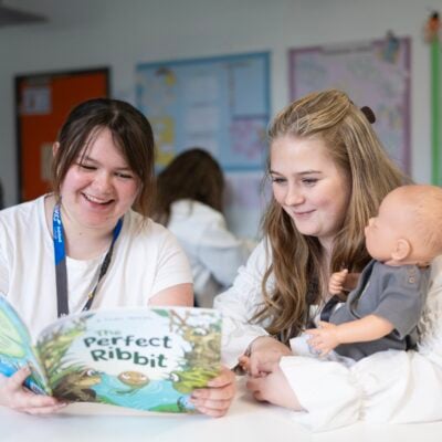 two students reading a book at a table to a doll.