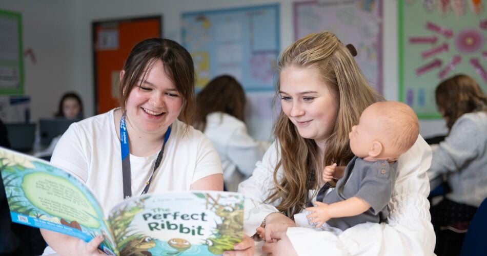 two students reading a book at a table to a doll.