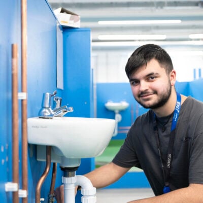 plumbing student looking at camera whilst working on a sink attached to a blue wall
