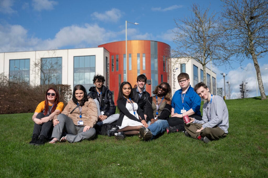 Group of students sitting on the grass smiling at the camera, the college is in the background