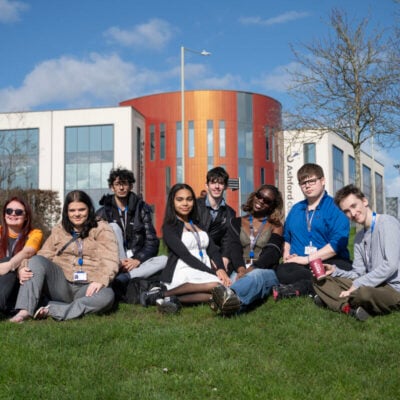 Group of students sitting on the grass smiling at the camera, the college is in the background