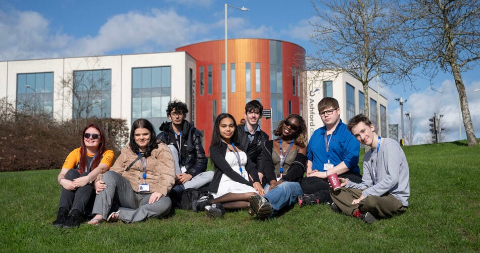 Group of students sitting on the grass smiling at the camera, the college is in the background