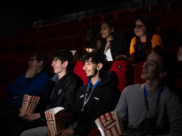 Rows of students sitting on red seats in a cinema screen in the dark holding popcorn. They are smiling.