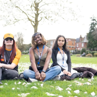 three students looking and smiling at the camera sitting on grass outside the college.