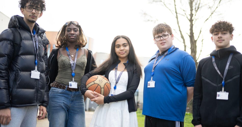 Five students looking at the camera standing outside the college. The student in the middle is holding a basketball.