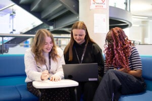 three students sitting at a desk, looking at a laptop. They are sitting on a dark blue sofa inside college.