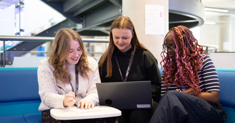 three students sitting at a desk, looking at a laptop. They are sitting on a dark blue sofa inside college.