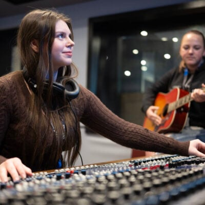 student using control music board and another student in the background playing a guitar