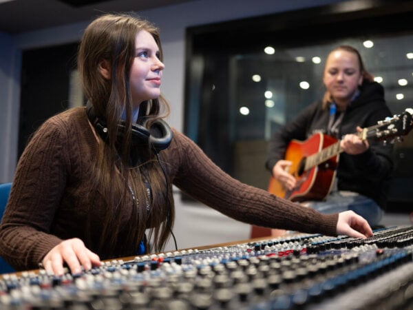 student using control music board and another student in the background playing a guitar