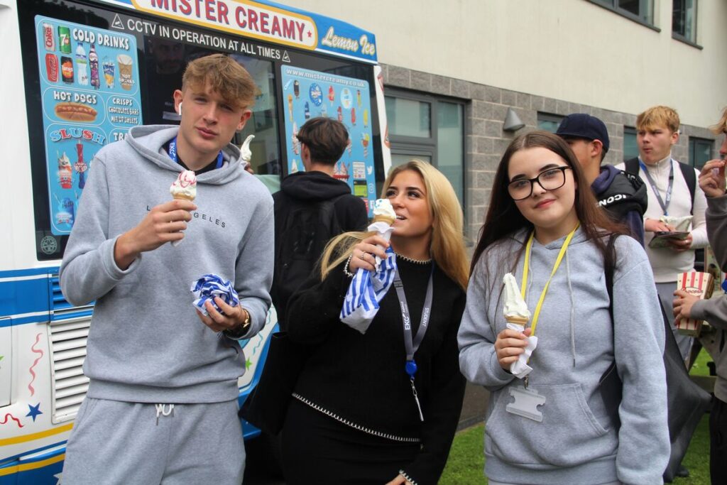Three students holding Mr Whippy ice creams and standing in front of an ice-cream van.