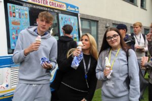 Three students holding Mr Whippy ice creams and standing in front of an ice-cream van.