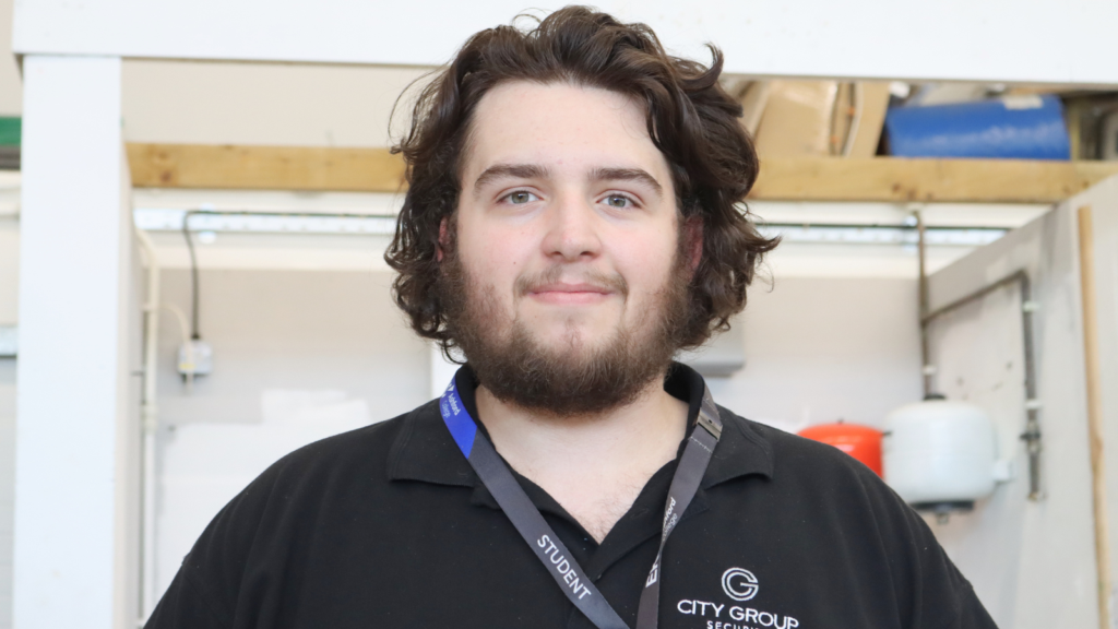 Joshua, student, smiling at the camera. He has dark brown wavy hair, a beard and is wearing a black polo shirt.