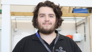 Joshua, student, smiling at the camera. He has dark brown wavy hair, a beard and is wearing a black polo shirt.