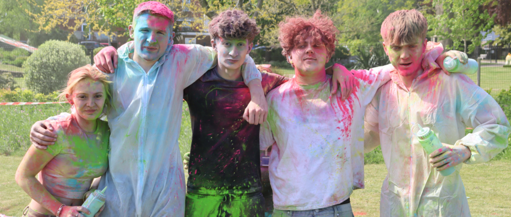 Four students covered in paint standing outside and looking at the camera, they are covered in pink, green and blue paint and have their arms around each other.