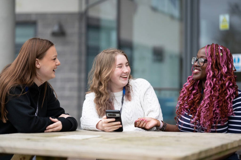 three students sitting on a picnic bench outside talking and laughing with each other.