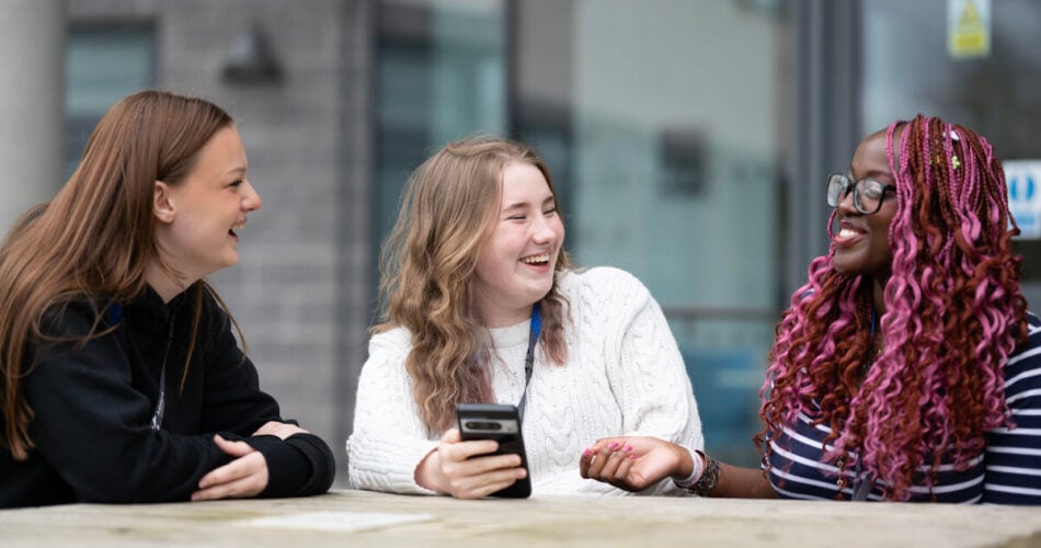 three students sitting on a picnic bench outside talking and laughing with each other.