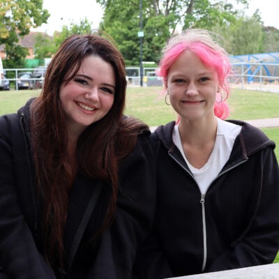 Two students sitting outside smiling at the camera. Both are wearing black hoodies. One has long brown hair and the other has pink hair in a ponytail.