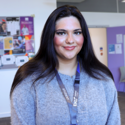 Student is smiling at the camera. She is standing in a classroom. She is wearing a light grey jumper and has long black hair.