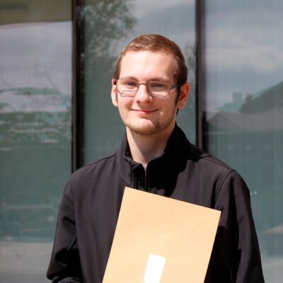 Student is wearing glasses and has short brown hair and is wearing a black shirt. He is standing outside and is holding a brown envelope.