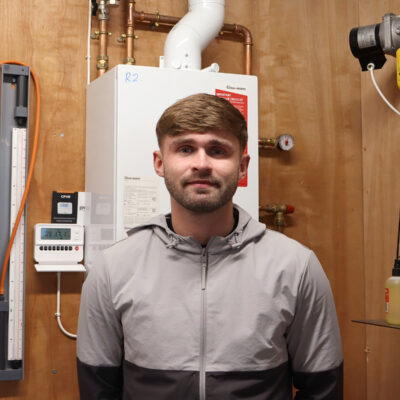 Student smiling at camera he has short brown hair and is wearing a grey jacket. He is standing in front of a boiler.
