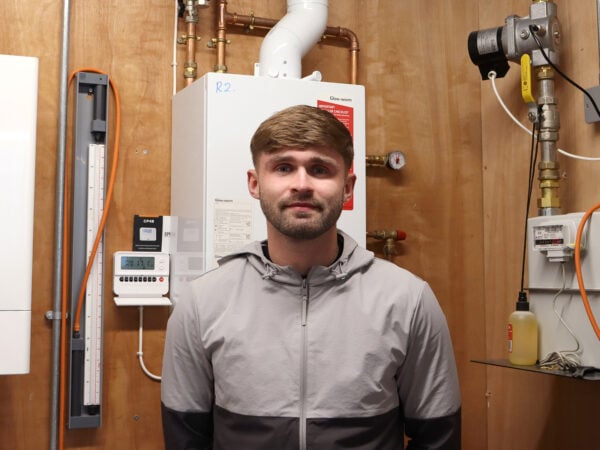Student smiling at camera he has short brown hair and is wearing a grey jacket. He is standing in front of a boiler.