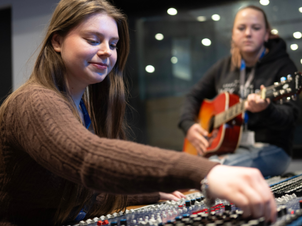 student using control music board and another student in the background playing a guitar