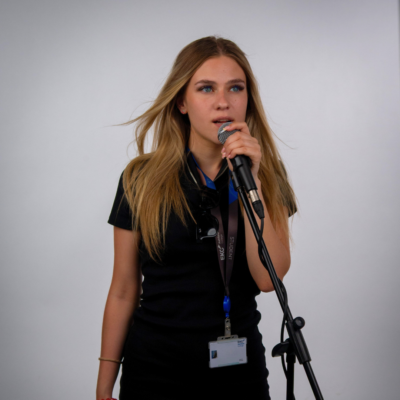 Student singing into microphone, she has long blonde straight hair and is wearing a black dress.