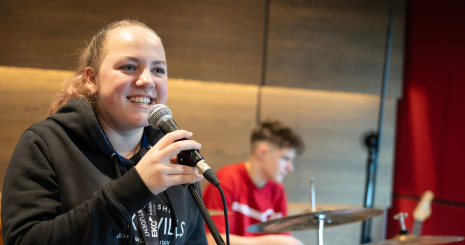 Two students in a music studio, student in front is holding a mic and smiling wearing a grey hoodie. Student in the background is playing the drums wearing a red top.