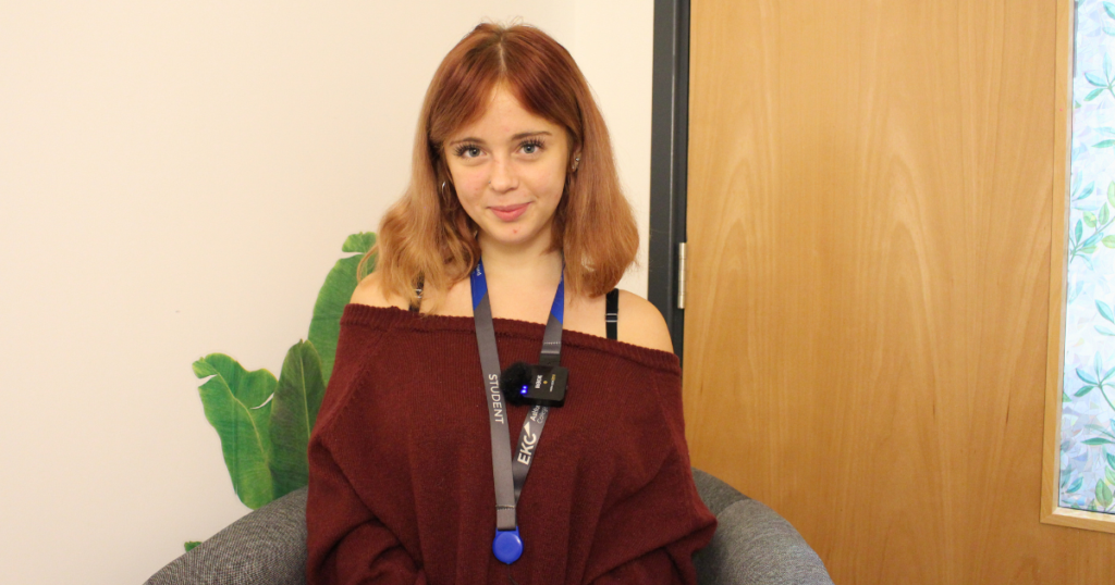 Student sitting on a grey chair, she is wearing a burgundy top and has shoulder length copper hair. She is smiling at the camera.