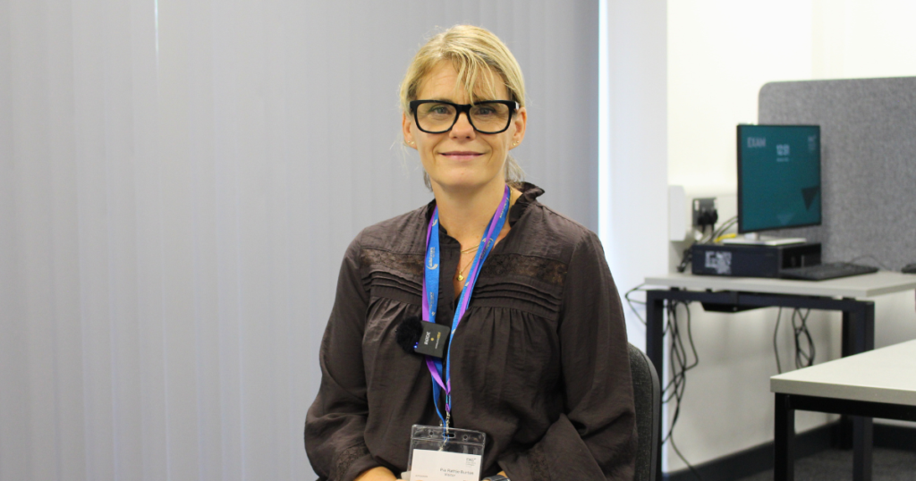 Guest speaker sitting on a chair looking and smiling at the camera, she is wearing glasses and has her hair tied up. She is wearing a dark grey blouse.