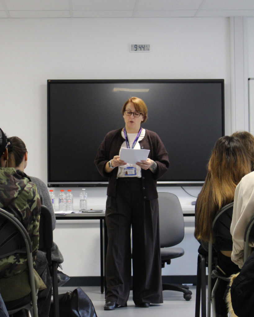 Guest speaker reading from a piece of paper, she is wearing black glasses and wearing a black cardigan and trousers, she has a brown bob haircut.