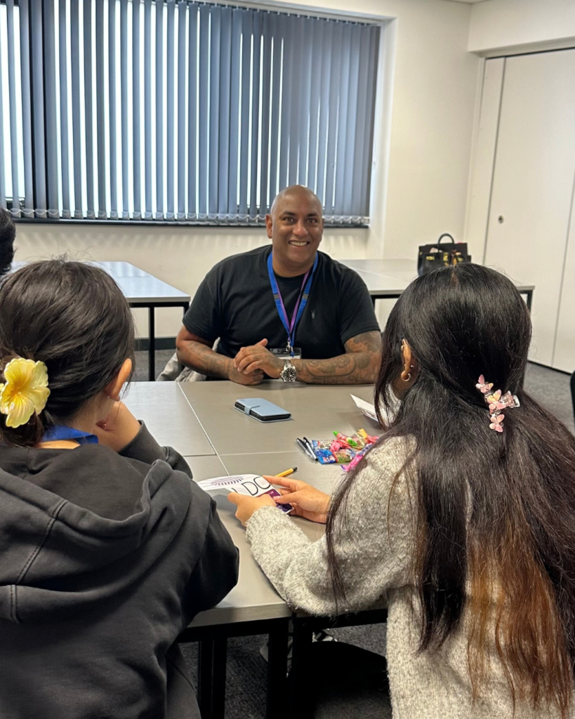 Guest speaker talking to students at a table in a classroom. He is smiling, wearing a black t-shirt, he is bald.