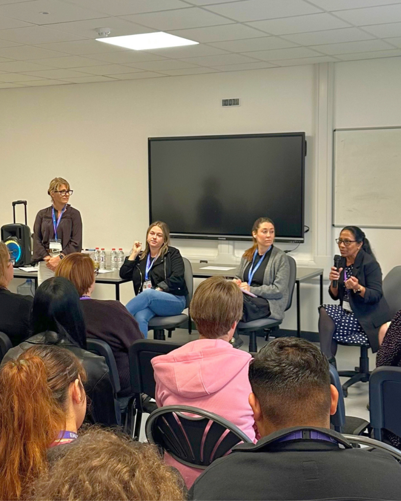 Three guest speakers sitting on chairs looking out to a room of students. One is holding a mic and speaking, she is wearing glasses and has black hair tied back into a ponytail talking to the room.
