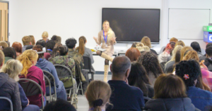 Woman with long blonde hair talking in front of a room of students.