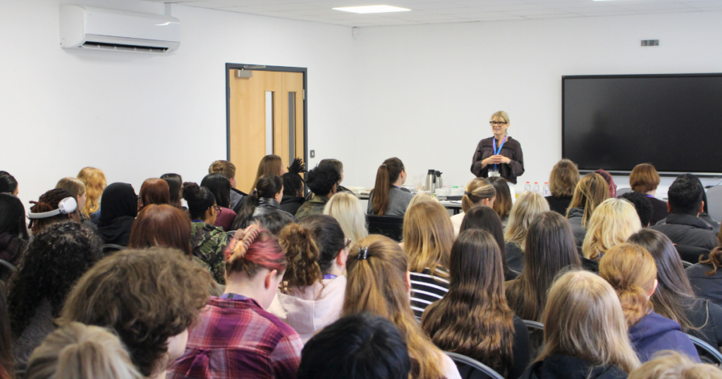 Woman with blonde hair wearing glasses talking in front of a room of students.