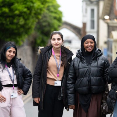 four students smiling at the camera outside on a street. They are all wearing black coats.