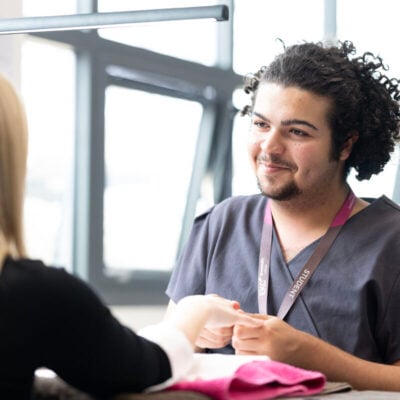 student sitting at a table, wearing a grey uniform, they are smiling at a client who is getting her nails done.