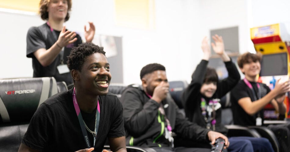 Five students playing video games and smiling, clapping and laughing together.