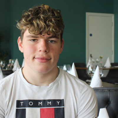 Student sitting on a chair in a restaurant. He is smiling at the camera and has mousey brown wavy hair, he is wearing a white t-shirt.